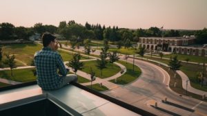 Man in a blue and white checked shirt looks out over a college campus. 