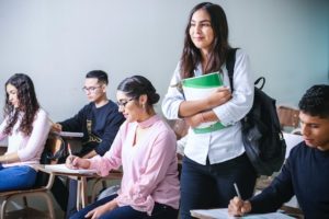 A young woman walks into a college classroom holding books. 